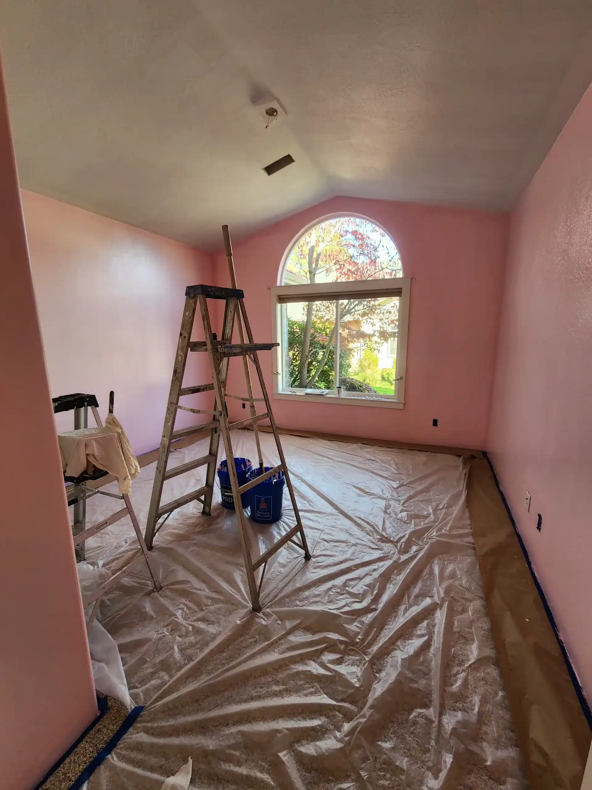Interior residential painting Vaulted bedroom with pink walls being repainted, ladder and drop cloths staged under an arched window