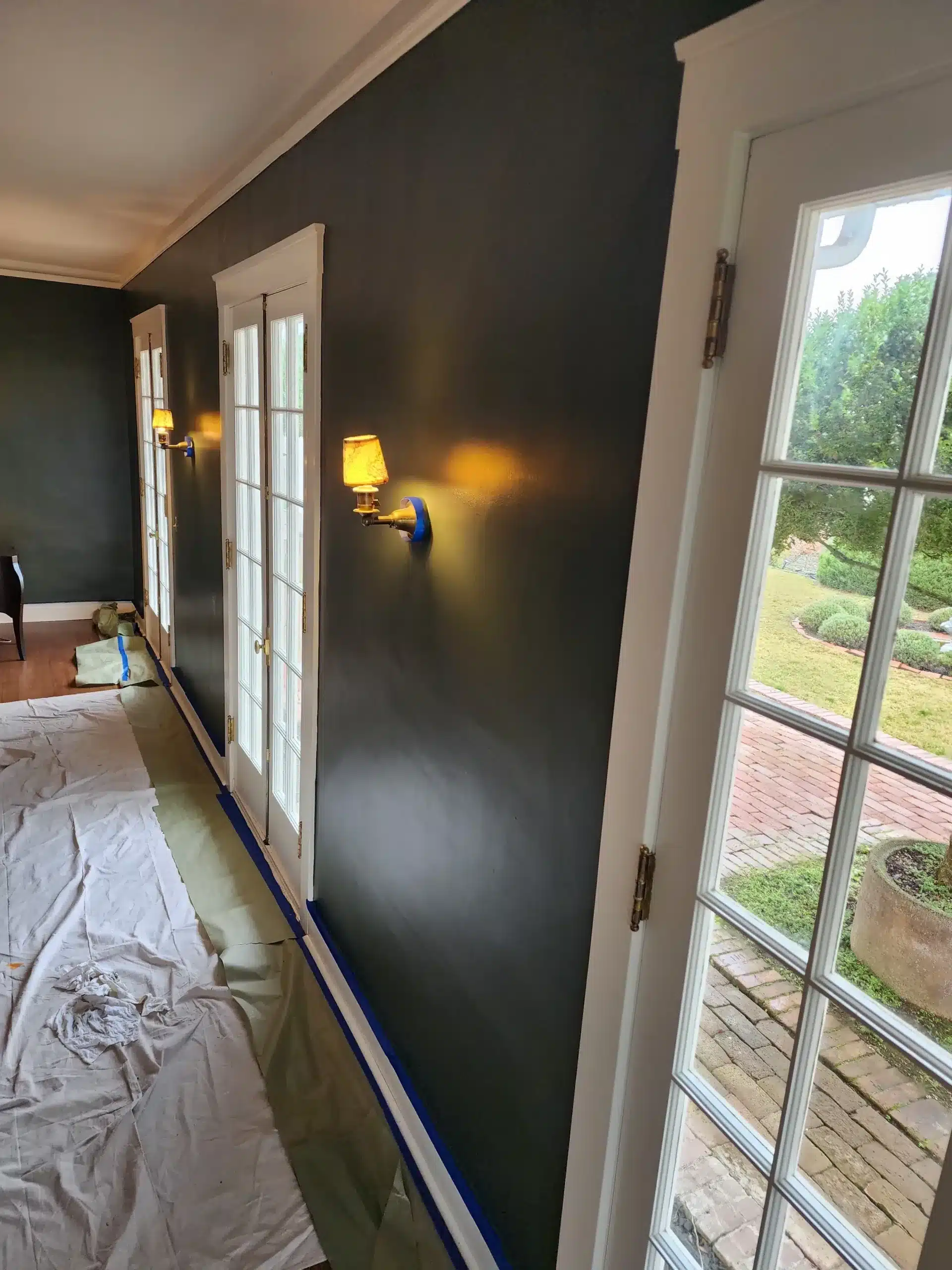 Interior residential painting Dark green interior walls mid-project with drop cloths and blue tape along a row of French doors overlooking a brick patio