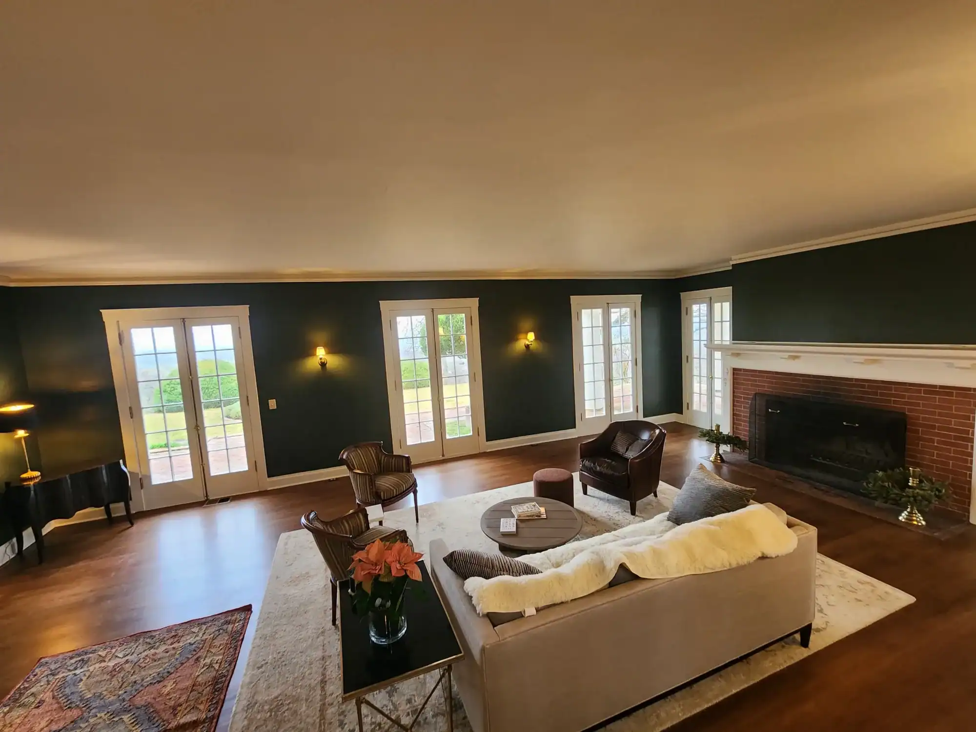 Interior residential painting Furnished living room with freshly painted dark green walls, white crown molding, brick fireplace, and a row of French doors overlooking a garden