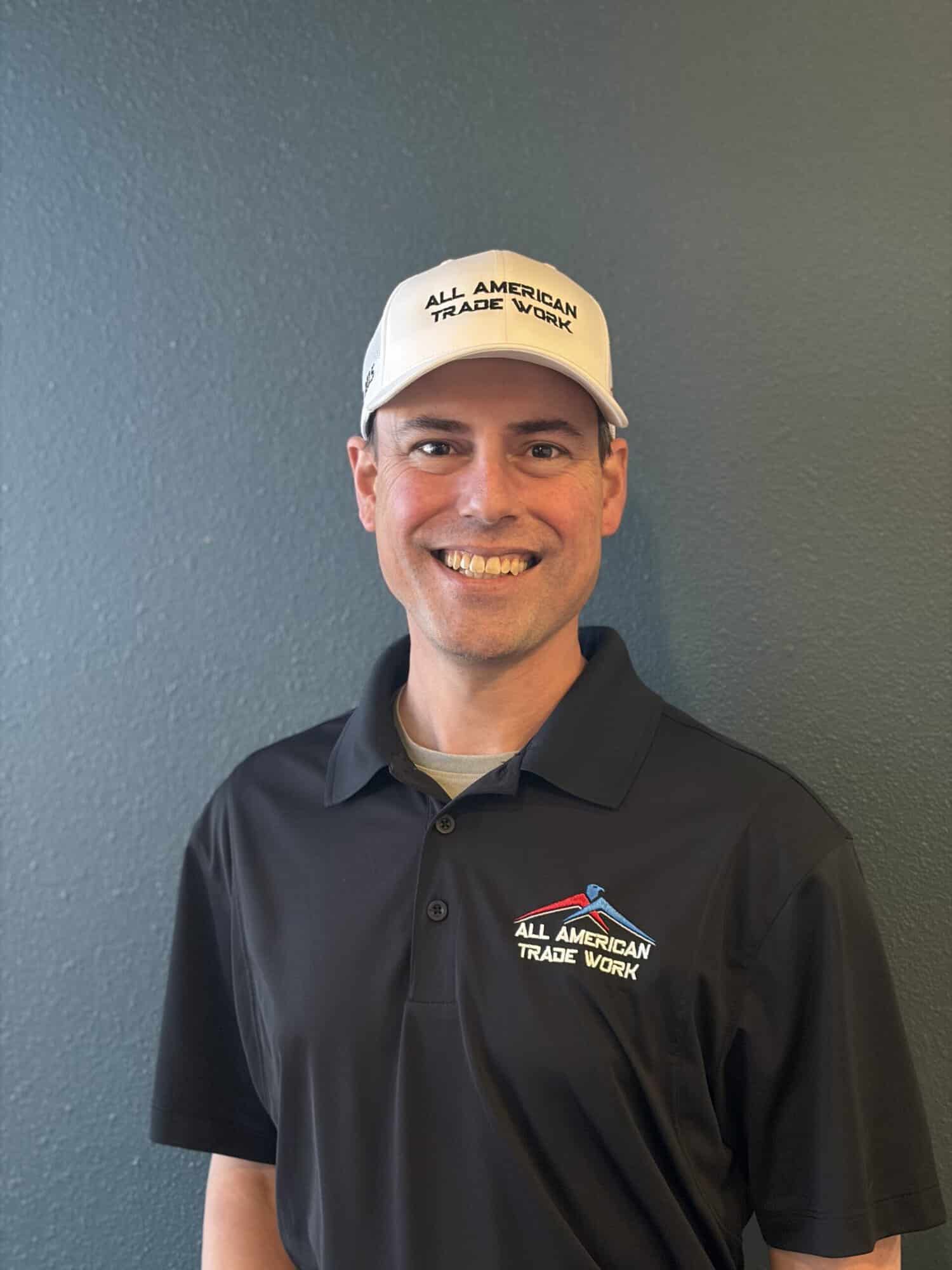 Painting Company Estimator Portrait Man wearing black polo shirt and white cap with All American Trade Work branding against gray painted wall